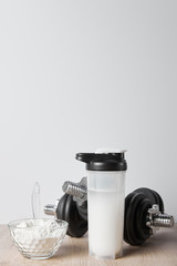 Obraz na płótnie Canvas sports bottle with protein shake near black dumbbells and glass bowl with protein shake isolated on grey