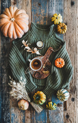 Autumn outfit layout. Flat-lay of dark green knitted sweater, blue jeans, hat, decorative pumpkins, candle, accessories and tea in cup on board over wooden background, top view. Fall apparel and mood