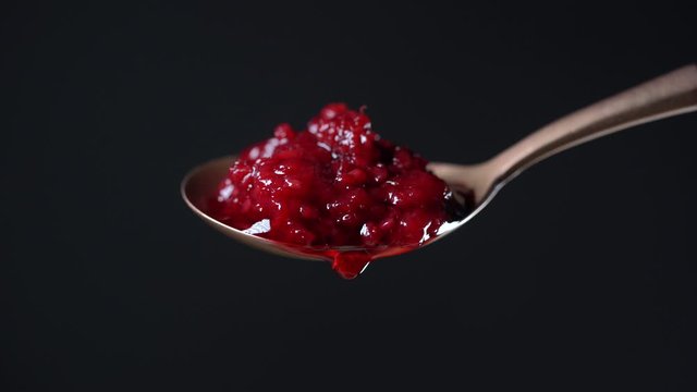 Spoon with red raspberry jam and one drop. Red raspberry jam dripping and flowing down from the metal spoon on black background, close up. Brass spoon with homemade red raspberry jam