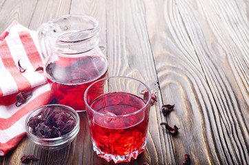 Closeup view at glass of hibiscus ice tea and jug on wooden table background