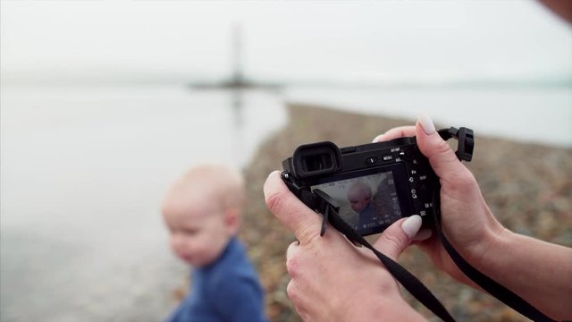 Camera in female hands. Mother is taking photos of her son who is sitting on pebble near water and playing with stones