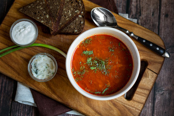 Borscht soup in a white bowl on the dark rustic wooden table with slices of dark bread, green onion, sour cream 