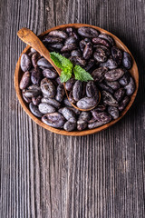 Whole dry cocoa beans in wooden bowl spoon mint on old rustic background.