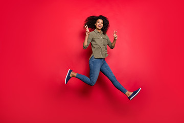 Full length body size photo of cheerful positive curly wavy brunette funky girlfriend running jumping showing v-sign wearing jeans denim shirt smiling toothily isolated vivid color background