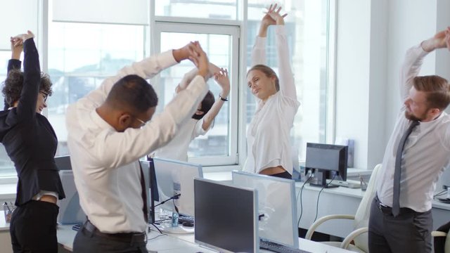 Medium shot of five businesspeople standing at their desks, doing stretching exercises together and then sitting down at desks