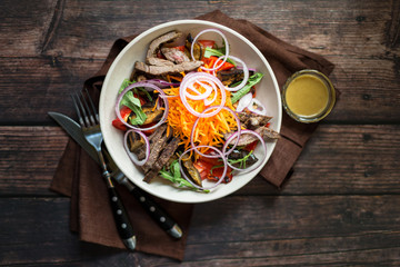 Salad with grilled beef, carrot, red onion and roasted vegetables with dressing on dark rustic wooden table