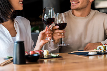 Cropped view of happy man and cheerful woman clinking glasses with red wine near sushi