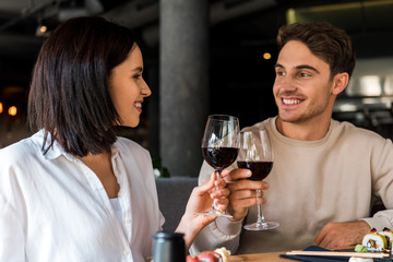happy man and cheerful woman clinking glasses with red wine near sushi