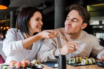 Fotobehang selective focus of happy woman holding chopsticks with tasty sushi near cheerful man in restaurant © LIGHTFIELD STUDIOS