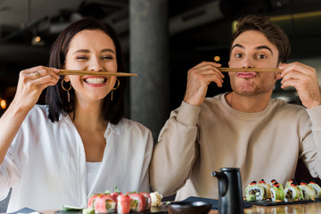 handsome man holding chopsticks near face while sitting with cheerful girl in sushi bar