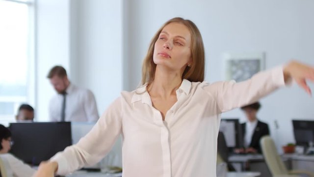 Medium shot of blonde businesswoman stretching her neck and then starting meditation with her eyes closed while other people working on background