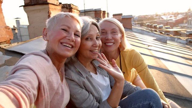 Point of view of three multiethnic female friends sitting together on roof and making selfie