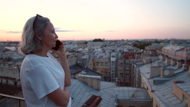 Medium shot of mixed-race elderly woman wearing white t-shirt standing on beautiful roof in city and talking on phone