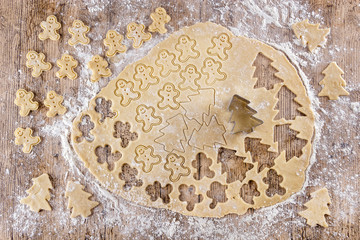 Top view of raw dough that is being cut to make cookies shaped like funny gingerbread dolls and Christmas trees on a floured wooden table. View from above. Recipes for children. Christmas desserts.