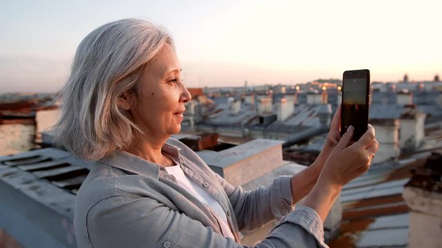 Medium shot of grey-haired pretty woman standing on building roof in historical center of city and making photos using smartphone