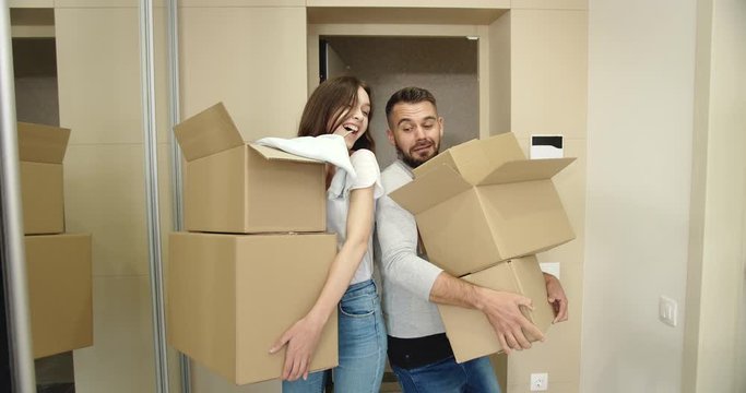 Happy young male and woman entering new apartment with cardboard boxes, laughing and having fun, new life beginning