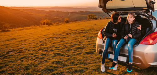 Mother and daughter sitting in car trunk on a hill and talking