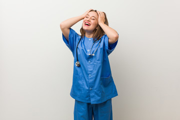 Young nurse woman against a white wall laughs joyfully keeping hands on head. Happiness concept.