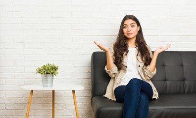 Young arab woman sitting on the sofa doubting and shrugging shoulders in questioning gesture.