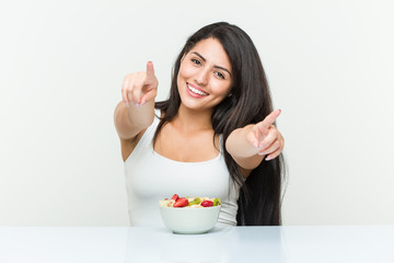 Young hispanic woman eating a fruit bowl cheerful smiles pointing to front.