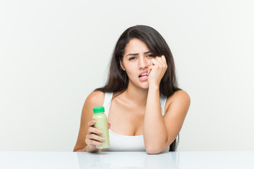 Young hispanic woman holding a vegetable drink biting fingernails, nervous and very anxious.