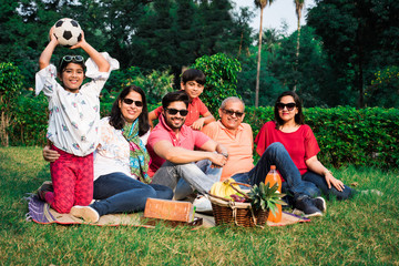 Indian Family enjoying Picnic - Multi generation of asian family sitting over lawn or green grass in park with fruit basket, mat and drinks. selective focus