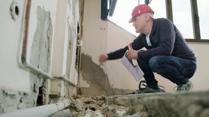 Technician working in construction site and checking wires and cables of electric power system near plumbing pipes. Man at work in apartment building, taking picture with camera phone