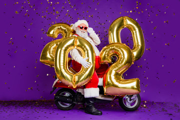Full body photo of fat santa man sitting on bike holding big air newyear 2020 numbers balloons confetti falling wear sun specs x-mas costume isolated purple background
