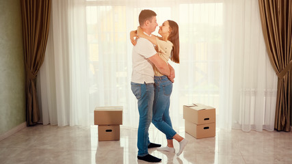 beautiful happy couple wearing blue jeans hugs and kisses standing in middle of new house lounge against big boxes, toned