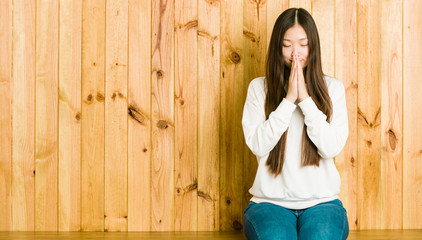 Fototapeta premium Young chinese woman sitting on a wooden place holding hands in pray near mouth, feels confident.