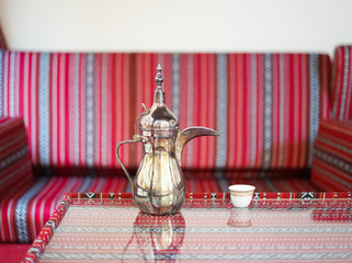 Arabic coffee. Coffee time served in a brass Arabic coffee jar with the blurred classic red Arabic seat in the background.