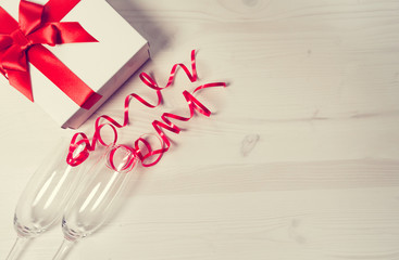 Two chrystal champagne glasses and a gift box tied with a red ribbon on a white painted wooden table.  Celebration background. Holiday and festive backdrop.