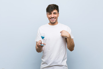 Young hispanic man holding a hourglass surprised pointing at himself, smiling broadly.