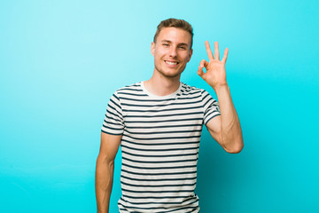 Young caucasian man against a blue wall cheerful and confident showing ok gesture.