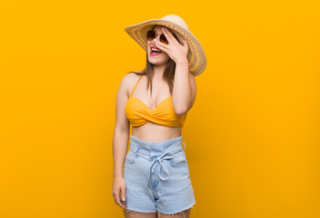 Young caucasian woman wearing a straw hat, summer look blink at the camera through fingers, embarrassed covering face.