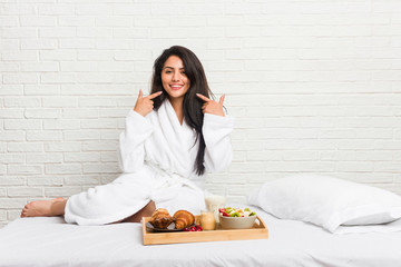 Young curvy woman taking a breakfast on the bed smiles, pointing fingers at mouth.