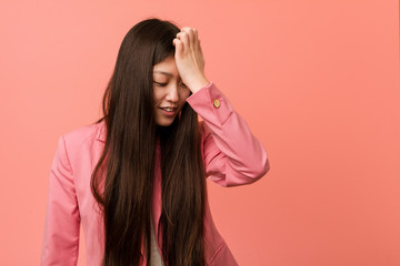 Young business chinese woman wearing pink suit forgetting something, slapping forehead with palm...