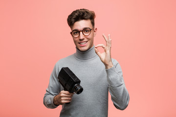 Young intellectual man holding a film camera cheerful and confident showing ok gesture.