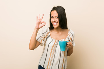 Young hispanic woman holding a cup cheerful and confident showing ok gesture.