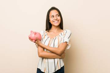 Young hispanic woman holding a piggy bank smiling confident with crossed arms.