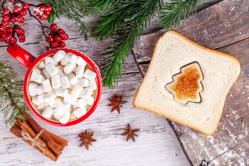 Cozy winter morning xmas food concept. Red cup with hot chocolate and marshmallows, toasted bread and fir tree shaped slices on wooden table in Christmas decorations. New Year breakfast.