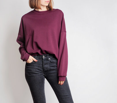 Young woman wearing simple outfit with oversized burgundy sweatshirt and black high-waisted jeans isolated on light grey background. Copy space