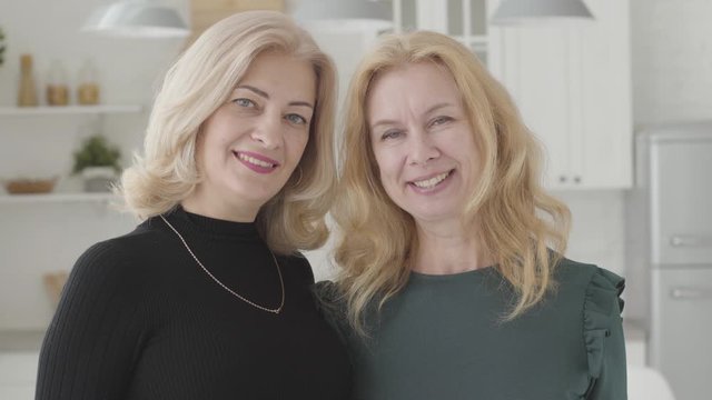 Close-up portrait of two beautiful mature Caucasian women looking at the camera and smiling. Positive attractive ladies standing in luxurious kitchen.
