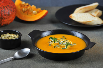 Pumpkin cream soup in a black plate. The soup is decorated with pumpkin seeds, cream and green sprouts. In the background are slices of toasted baguette. Close-up.