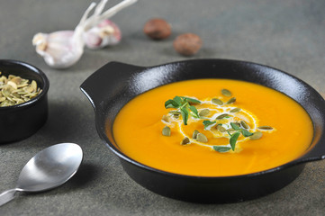 Pumpkin cream soup in a black plate. The soup is decorated with pumpkin seeds, cream and green sprouts. In the background are nutmeg, garlic and pumpkin seeds. Close-up.