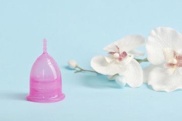 Obraz na płótnie Canvas Pink menstrual cup in the shape of a lily on a light background. Nearby is an orchid flower. The concept of cleanliness, comfort and freshness. Free space for text.