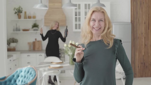 Rich mature Caucasian woman with a cigar scaterring money and smiling. Her positive friend dacing with wine and glasses at the background. Two wealthy senior ladies resting in the luxurious kitchen.
