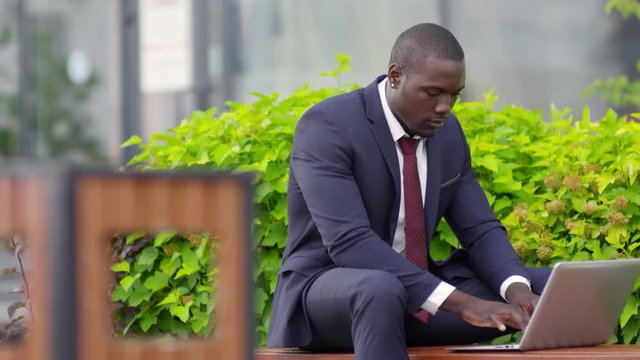 Tracking medium shot of young black man in business suit sitting on bench in park and working online using laptop computer