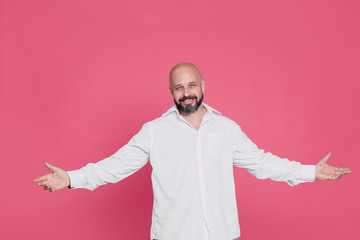Handsome serious middle-aged man in a white shirt smiles and spreads two hands in different directions on a pink background with copy space