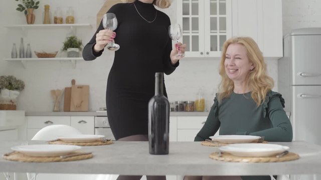 Two beautiful blonde Caucasian women sitting in the luxurious kitchen preparing to drink wine. Mature rich lady in elegant dress receiving guest at home.